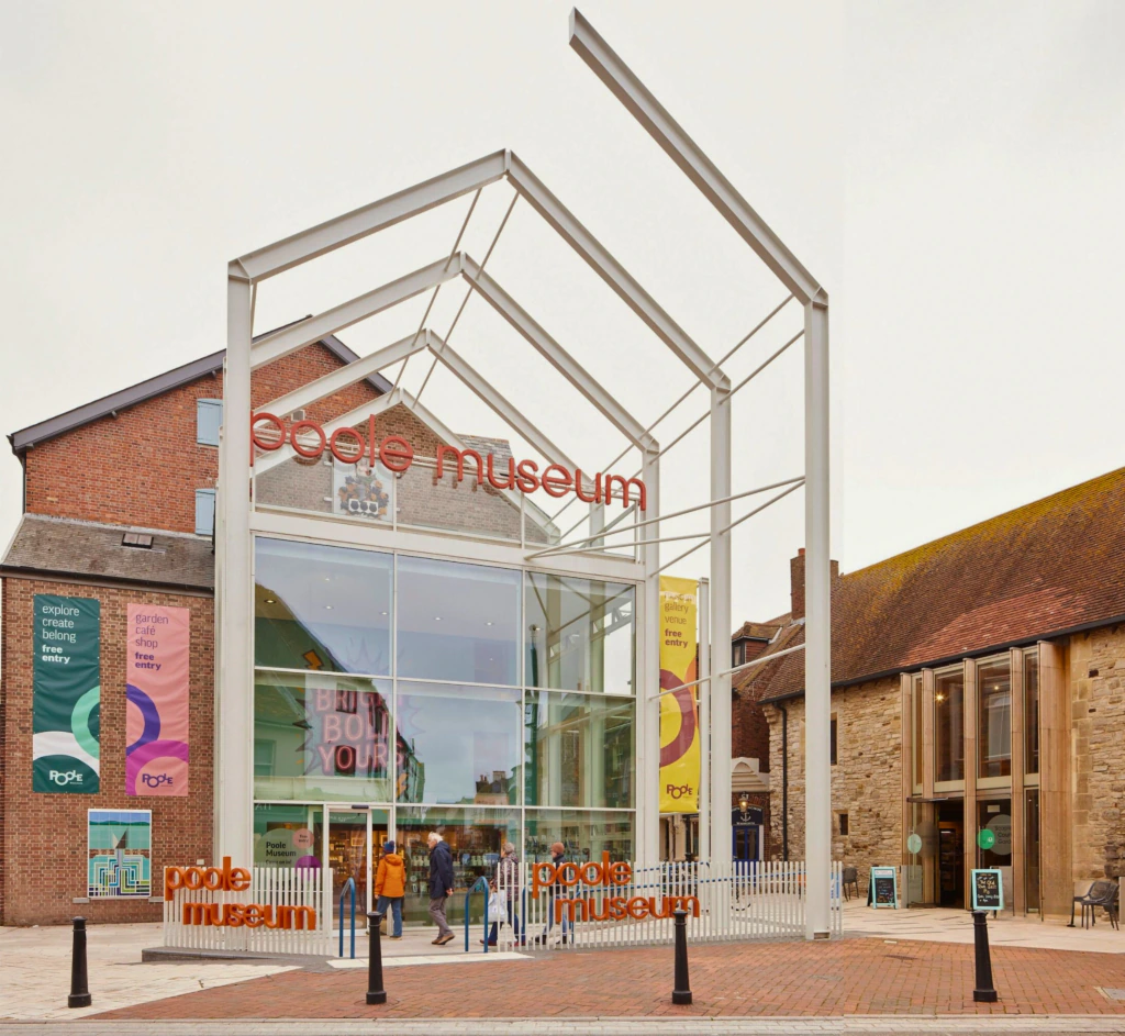 The image shows the modern glass entrance of the People’s Museum, framed by a white metal structure with the museum’s name in red letters. Colorful banners hang on both sides; brick buildings surround the entrance.