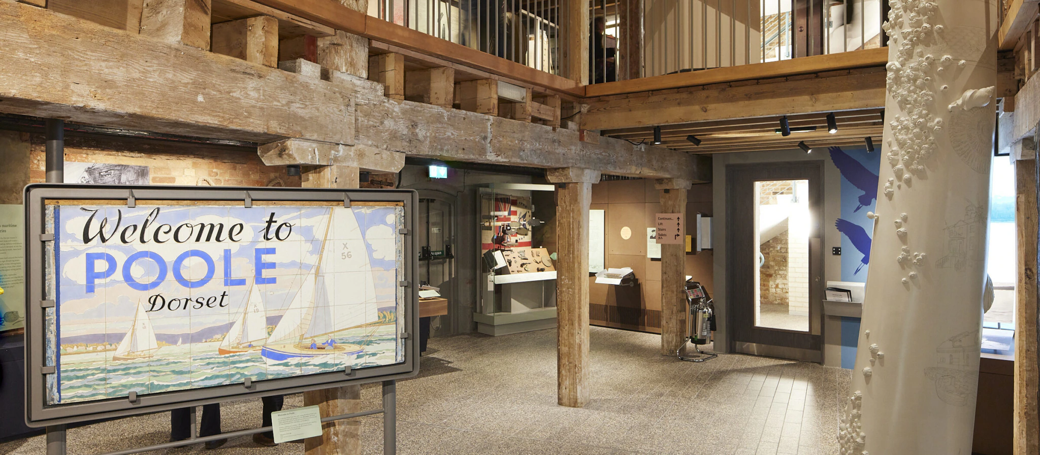 A museum interior with wooden beams, a two-story balcony, a tall decorative pillar, and a large sign reading Welcome to Poole Dorset featuring sailboats. Display cases and exhibits are visible in the background.