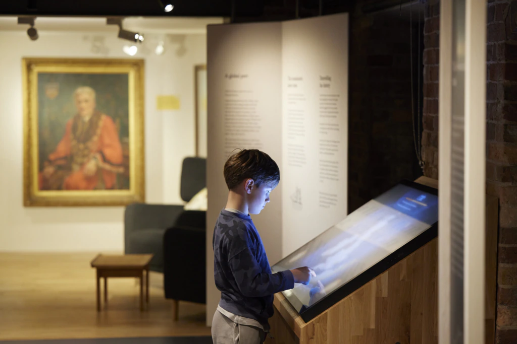 A young boy using a large interactive touchscreen display in a museum, with an illuminated portrait and informational panels visible in the softly lit background.