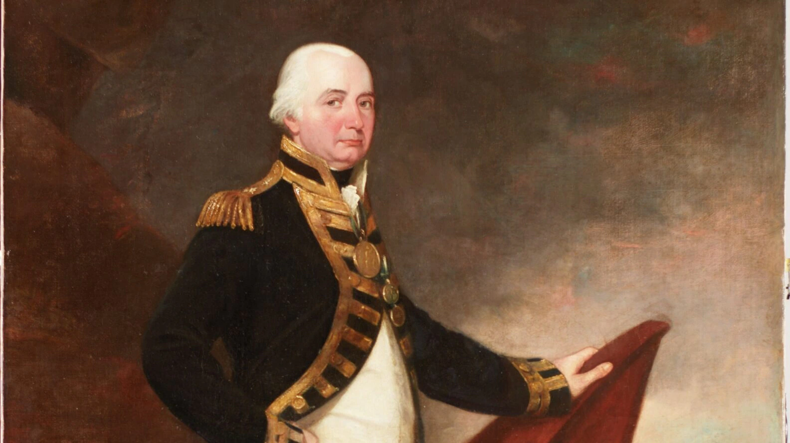 A man in an 18th-century naval officer’s uniform stands on rocky ground by the sea, one hand on a red cape or flag, with ships visible in the background under a cloudy sky.