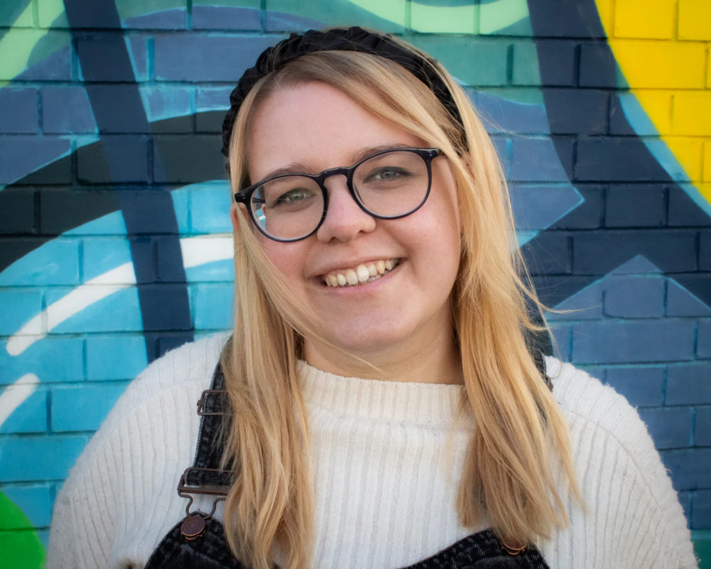 A person with long blonde hair, glasses, and a black headband smiles at the camera. They are wearing a white sweater and denim overalls, standing in front of a colorful, geometric mural on a brick wall.
