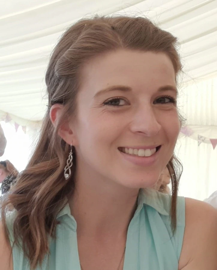 A woman with light brown hair and wavy curls, wearing a sleeveless light blue top and silver drop earrings, smiles at the camera in a bright indoor setting.
