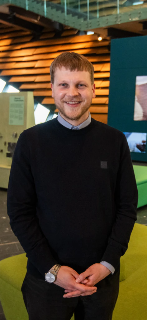 A man with short light brown hair and a beard, wearing a black sweater over a collared shirt, stands indoors smiling, with modern wooden and teal wall panels and green furniture in the background.