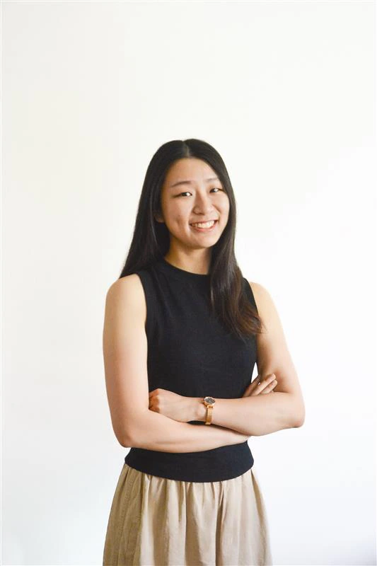 A woman with long dark hair, wearing a sleeveless black top and beige skirt, stands in front of a plain white background, smiling with her arms crossed.