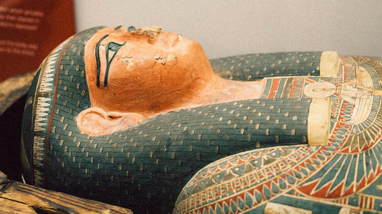 A close-up of an ancient Egyptian sarcophagus, decorated with colorful patterns and detailed facial features. The surface shows some chips and signs of age.