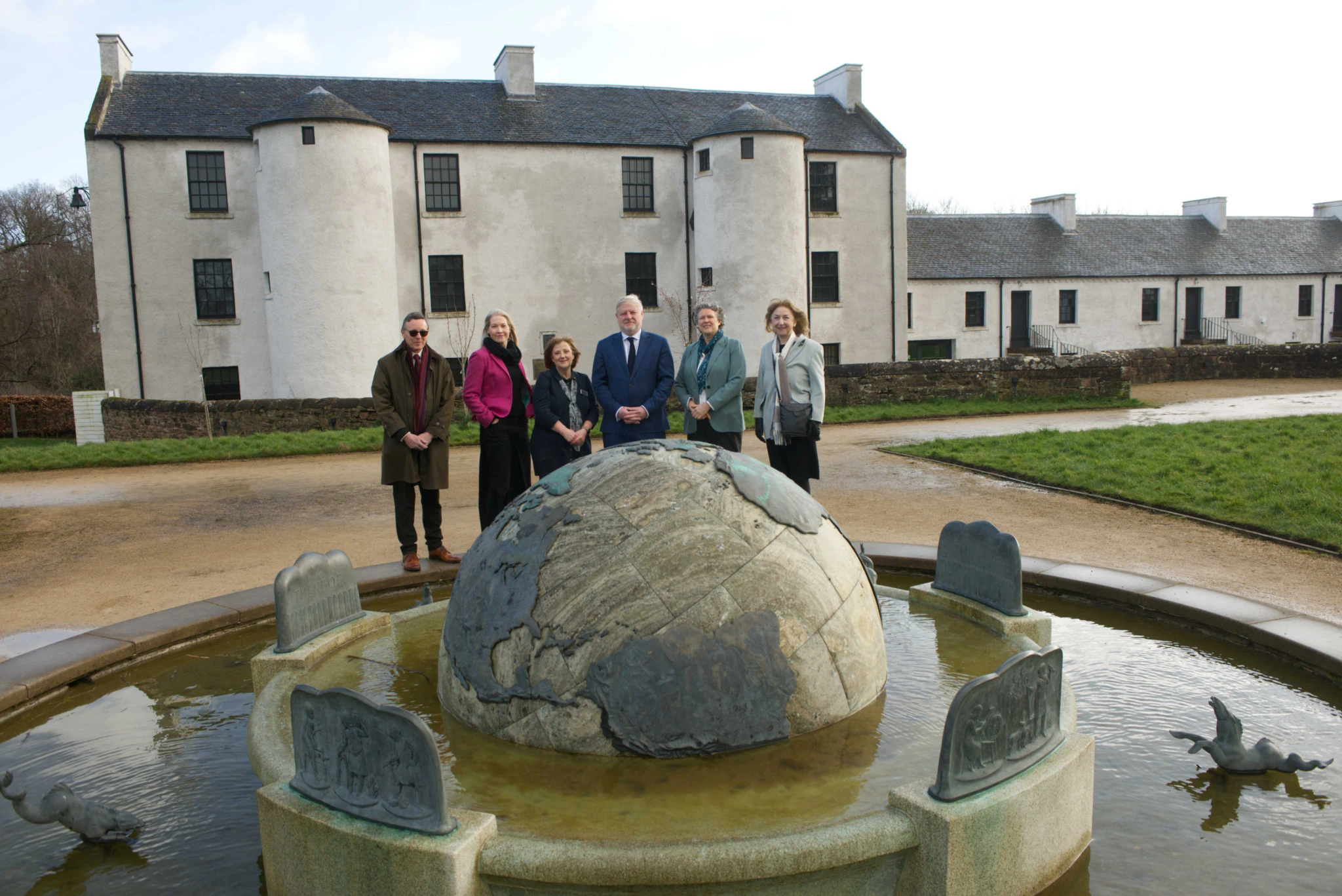 Six people stand behind a stone globe fountain with animal carvings, in front of a large white historic building with towers, on a cloudy day.