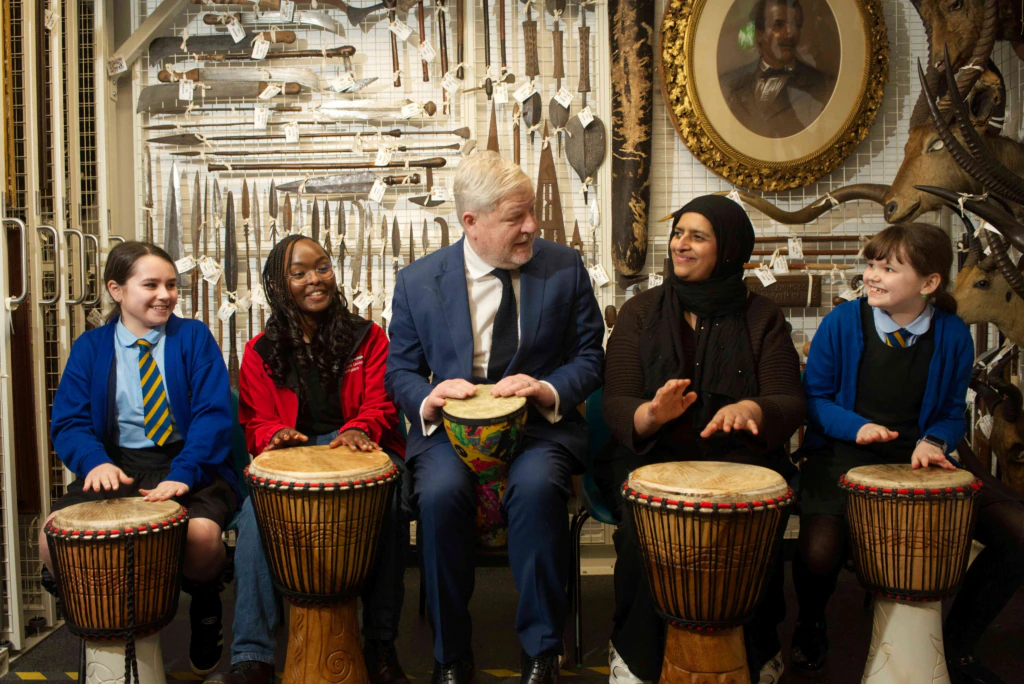 A group of five people, including three girls, a man in a suit, and a woman in a hijab, sit in a row playing hand drums in front of a wall displaying historical artifacts and a framed portrait.
