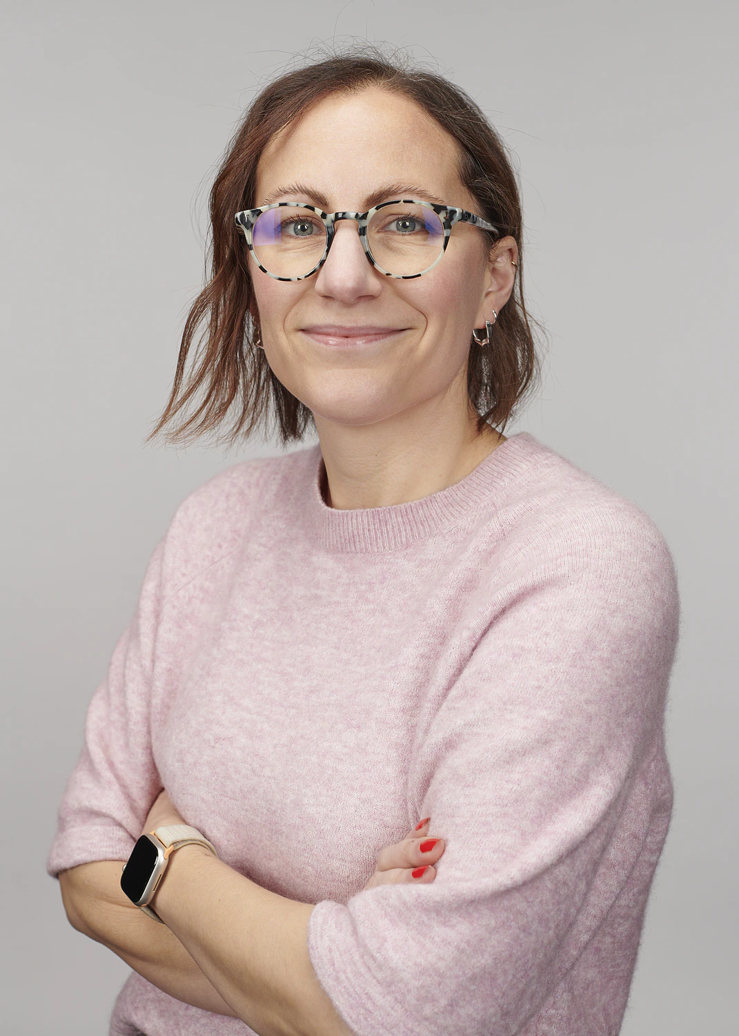 A woman with light brown hair wearing tortoiseshell glasses and a light pink sweater stands with arms crossed, smiling slightly. She has red nail polish, small hoop earrings, and an Apple Watch. The background is plain gray.