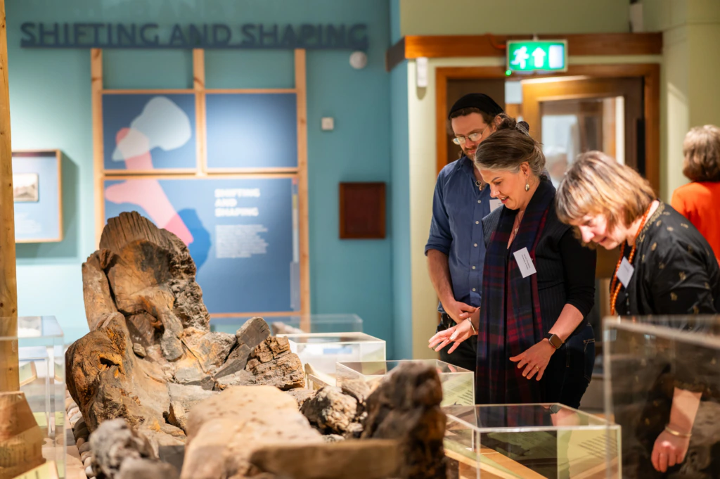 Three people look closely at a large, ancient wooden artifact displayed in a museum. Exhibition panels and glass display cases are visible in the background.