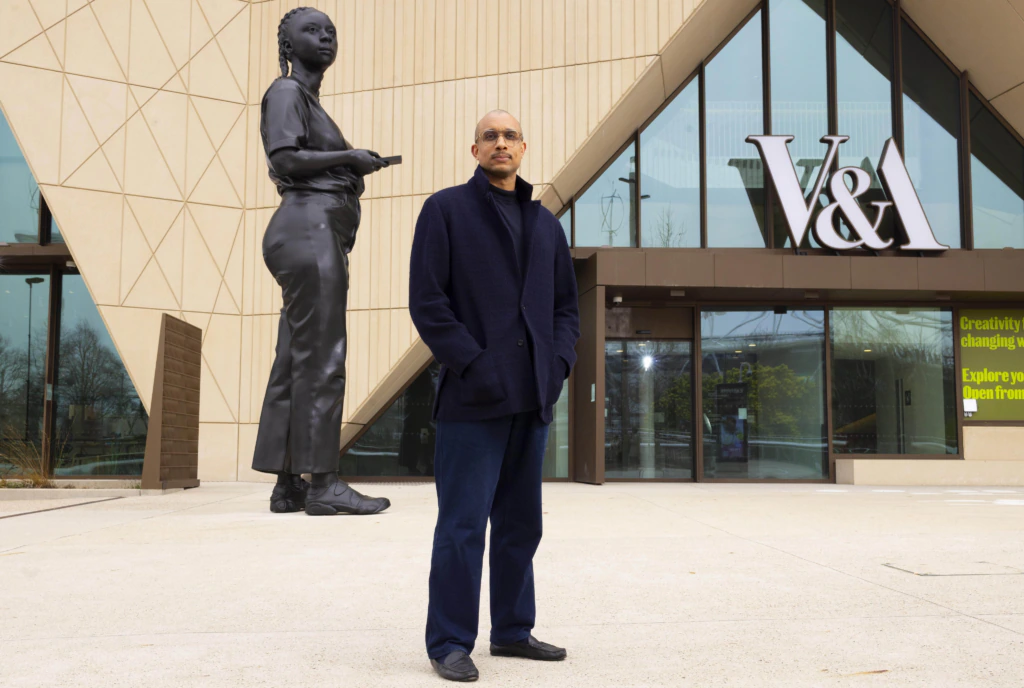 A person stands in front of the V&A building next to a large statue of a young woman, holding a phone. The building has modern architecture with glass doors and geometric patterns.