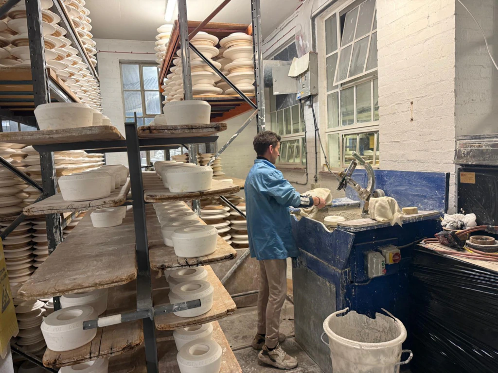 A person in a blue jacket shapes pottery on a machine in a workshop, surrounded by shelves filled with white ceramic bowls and plates. Bright light comes through large windows on the right.