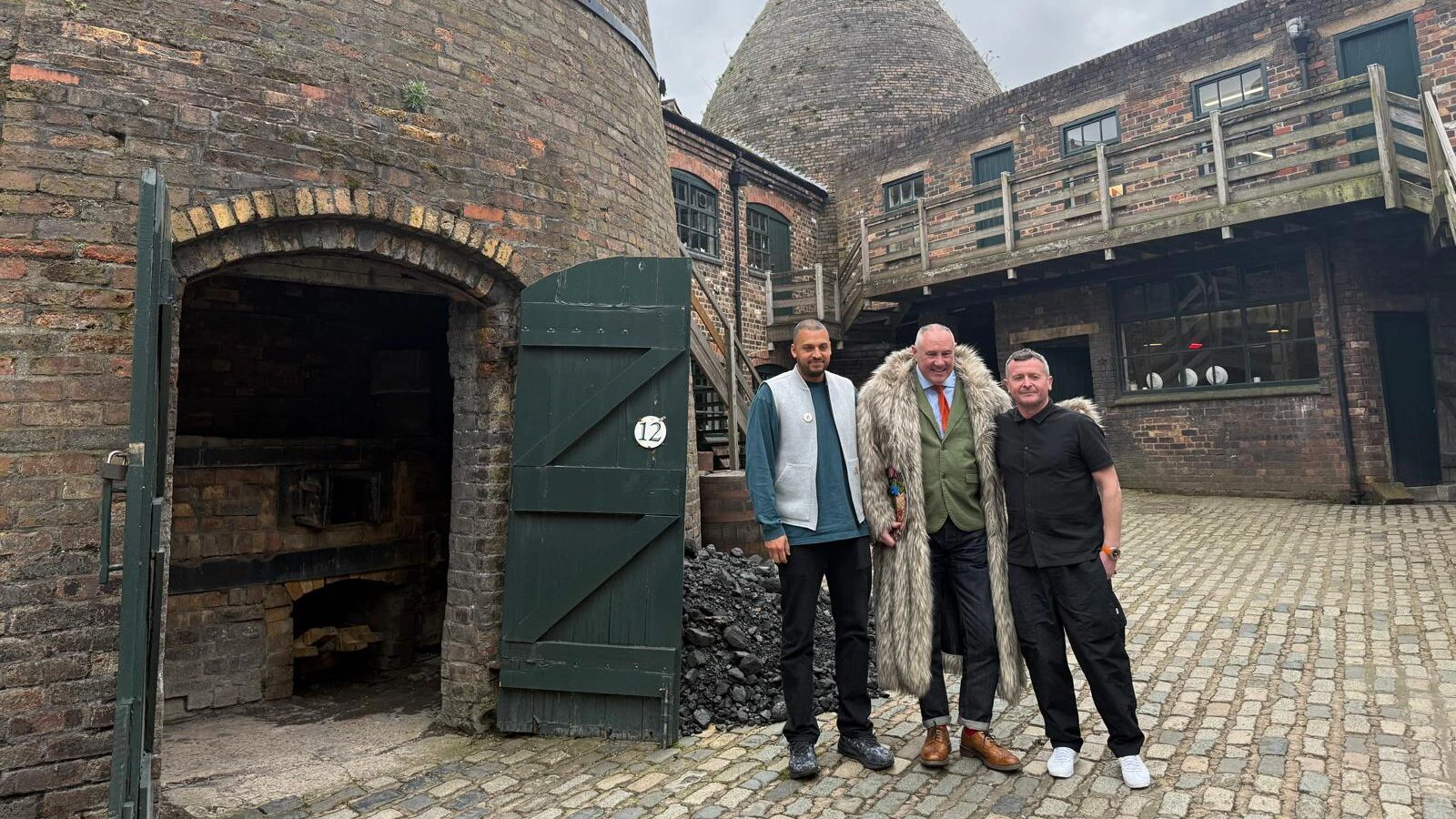 Three men stand together on a cobblestone courtyard outside old brick bottle kilns at an industrial heritage site, under a cloudy sky. One kiln door is open, revealing a dark interior.