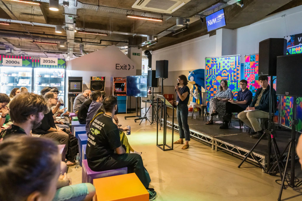 A speaker stands at a podium addressing an attentive audience at an indoor event, while three panelists sit on stage beside her. The room features colorful decorations and display cases in the background.
