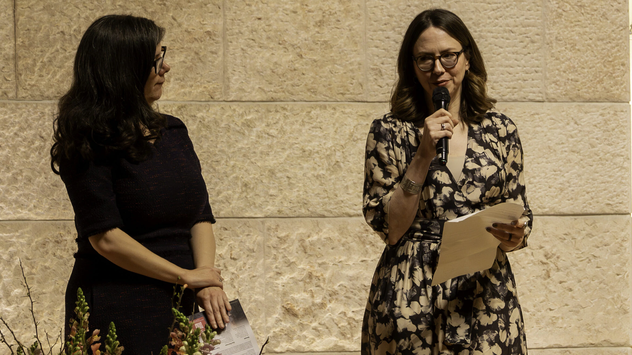 Two women stand indoors against a light stone wall. One, in a patterned dress, holds a microphone and papers while speaking. The other, in a dark dress, stands beside her holding papers. Flowers are visible in the foreground.