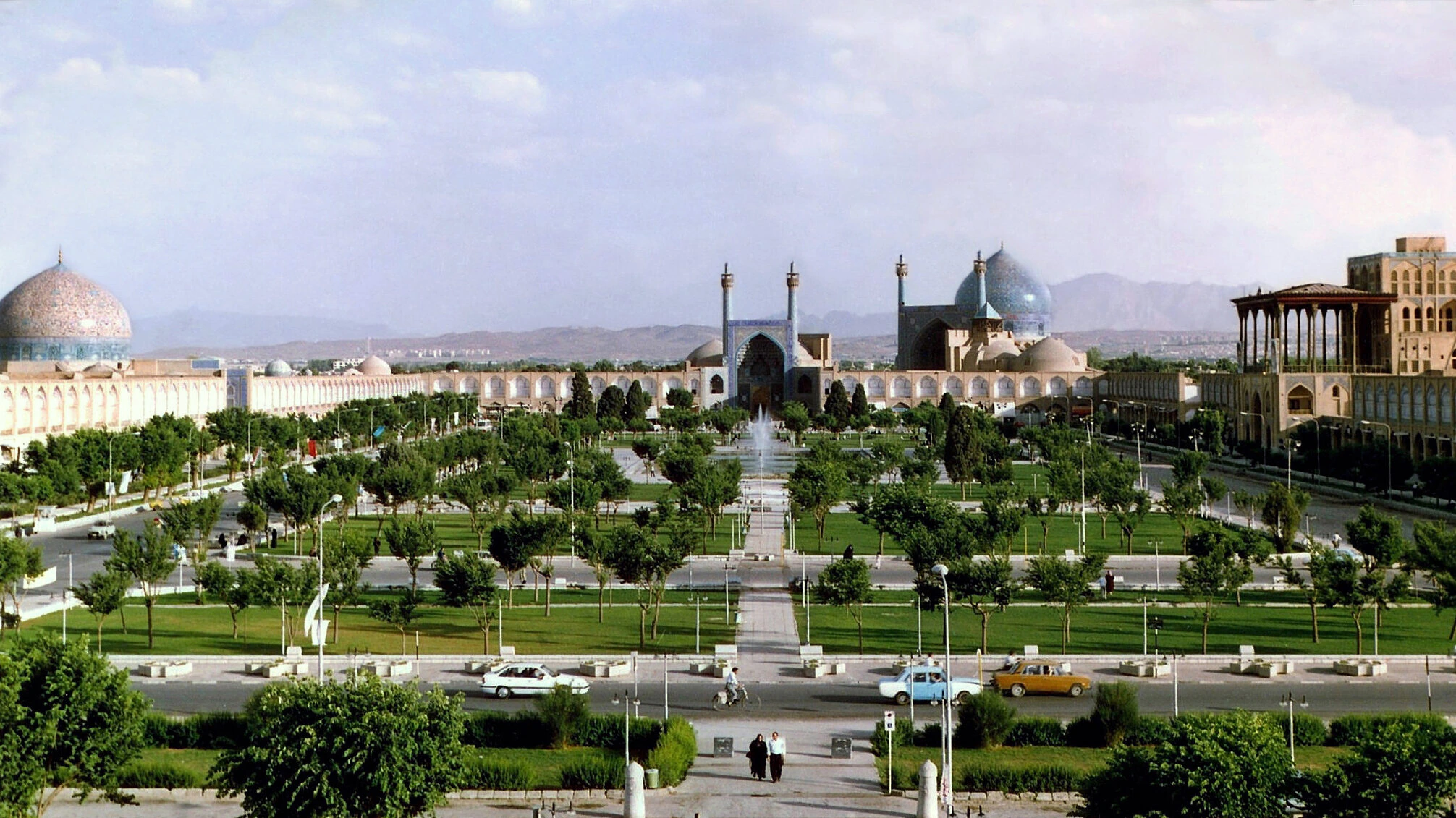 A panoramic view of Naqsh-e Jahan Square in Isfahan, Iran, showing lush gardens, historic mosques with blue domes, arched buildings, walkways, and people strolling on a sunny day.