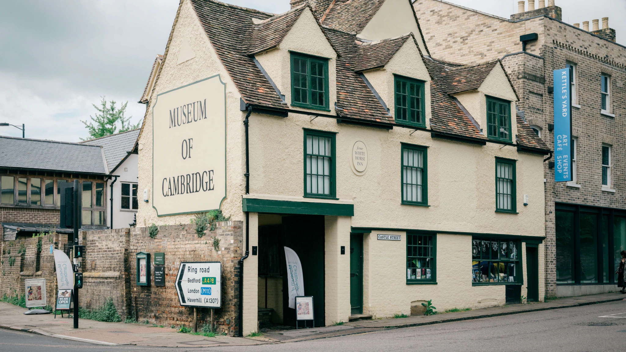 A historic cream-colored building with green trim houses the Museum of Cambridge. The building has a sloped roof, multiple chimneys, and signs at the front entrance. The street and nearby buildings are visible.