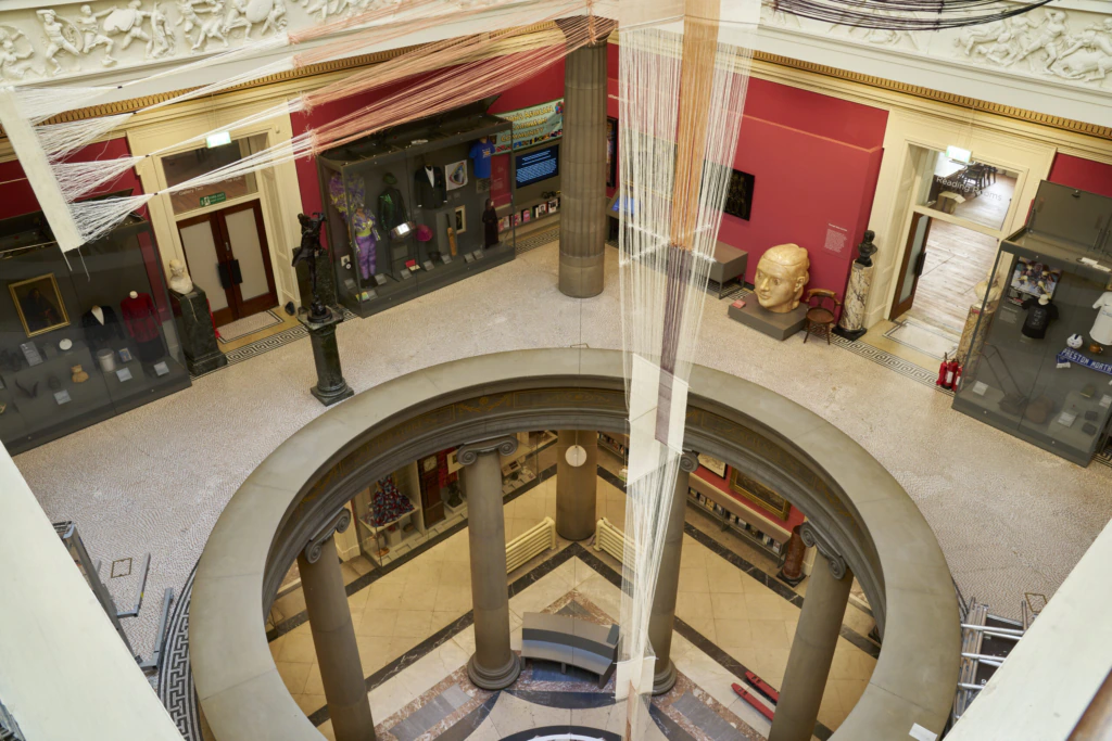 A view from above of a museum interior with a circular balcony, classical columns, display cases, red walls, and a large sculpture of a head on the floor. Artifacts and exhibits are visible around the room.