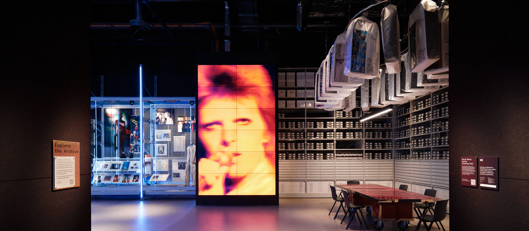 A modern museum exhibit features a large, illuminated image of David Bowie’s face, display cases with memorabilia, a table with chairs, and shelves filled with archive boxes under hanging photographs.