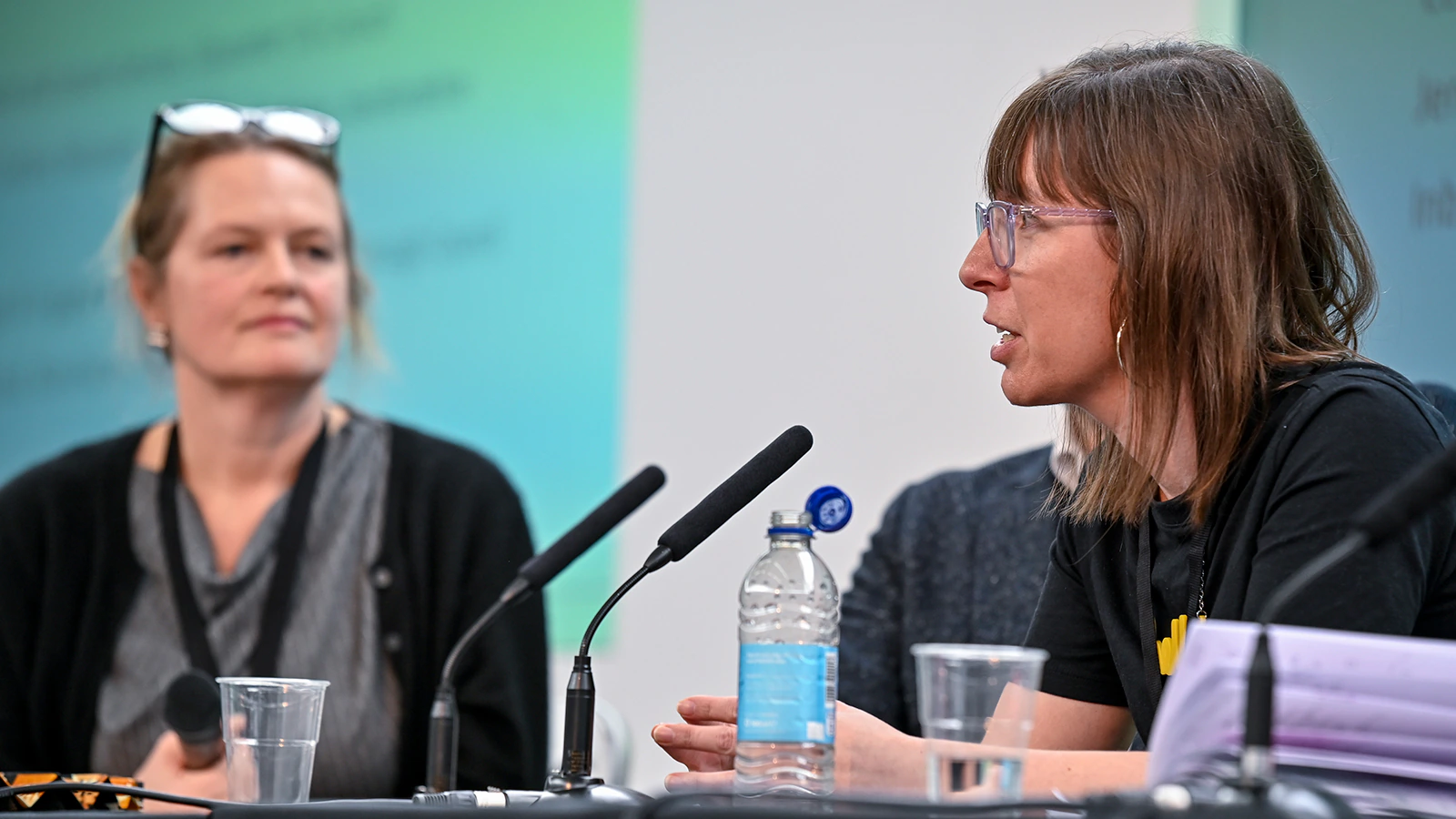 Two women sit at a table with microphones, engaged in discussion. One woman speaks while the other listens attentively. A water bottle, papers, and plastic cups are on the table in front of them