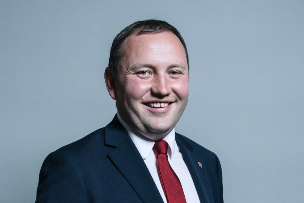 A man in a dark suit, white shirt, and red tie is smiling against a plain light gray background.
