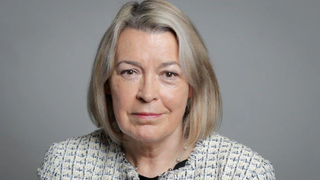 A middle-aged woman with straight, shoulder-length blonde hair, wearing a light-colored patterned jacket over a black top, looks directly at the camera against a plain gray background.