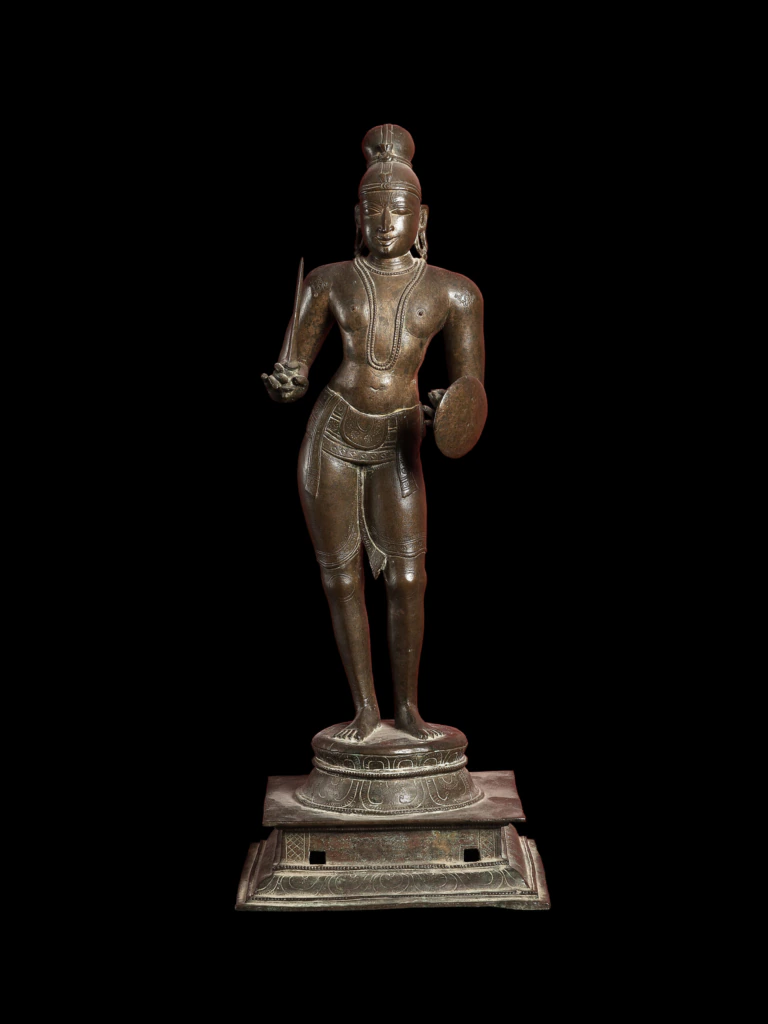 A bronze statue of a standing Hindu deity, holding a small disc and conch, adorned with jewelry and a headdress, displayed on a square pedestal against a black background.