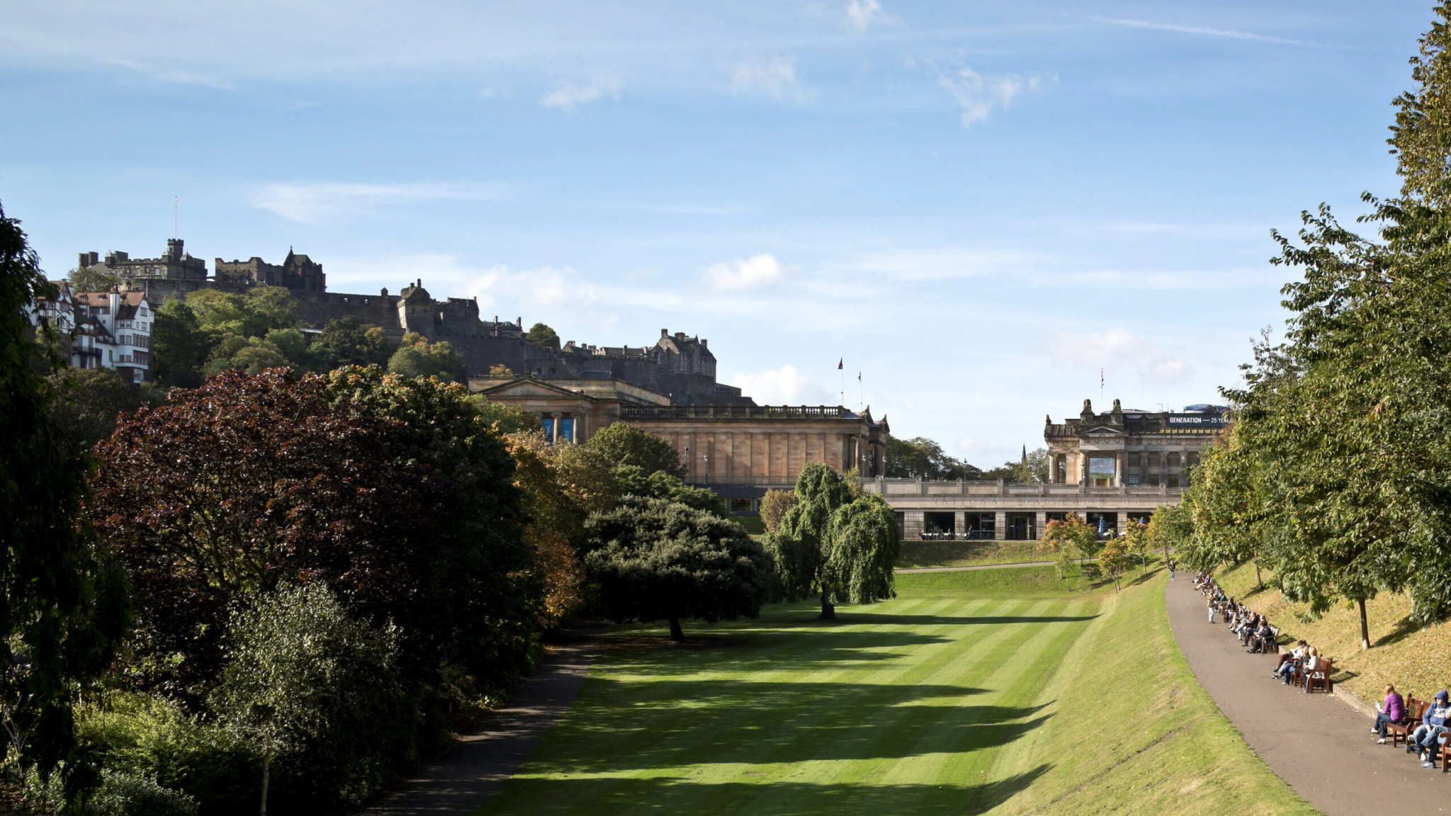 A wide grassy park with people sitting on benches along a path, bordered by trees. In the background, there are historic buildings and a large castle on a hill under a clear blue sky.