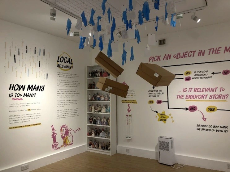 An art gallery room with interactive displays, hanging gloves, and floating cardboard boxes. Walls have colorful text, diagrams, and illustrations, with a shelf displaying numerous figurines in the corner.