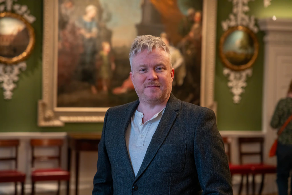 A man with short blond hair and a beard, wearing a gray blazer and light shirt, stands in an elegant room with green walls, ornate decor, and a large classical painting in the background.