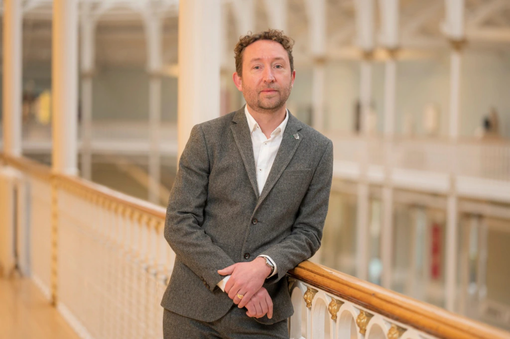 A man in a grey suit leans casually against a wooden and white railing in a bright, spacious indoor setting with high ceilings and blurred background details.