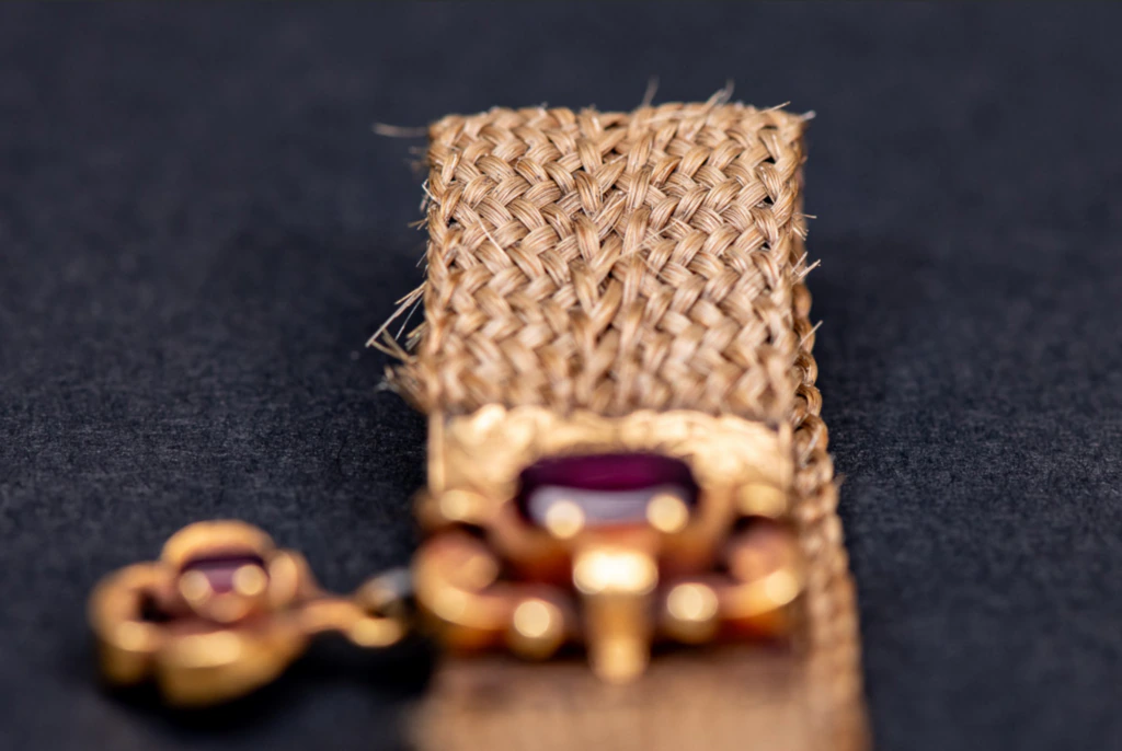 A close-up of a woven, tan-colored fabric strap with a decorative gold and maroon ornament attached, set against a dark background.