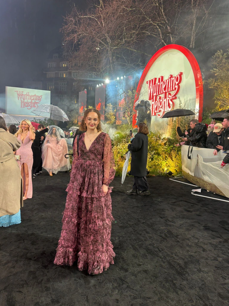A woman in a ruffled mauve gown stands on a black carpet at an outdoor event. Behind her, people gather near a large Wuthering Heights sign, with trees and lights creating a dramatic atmosphere.