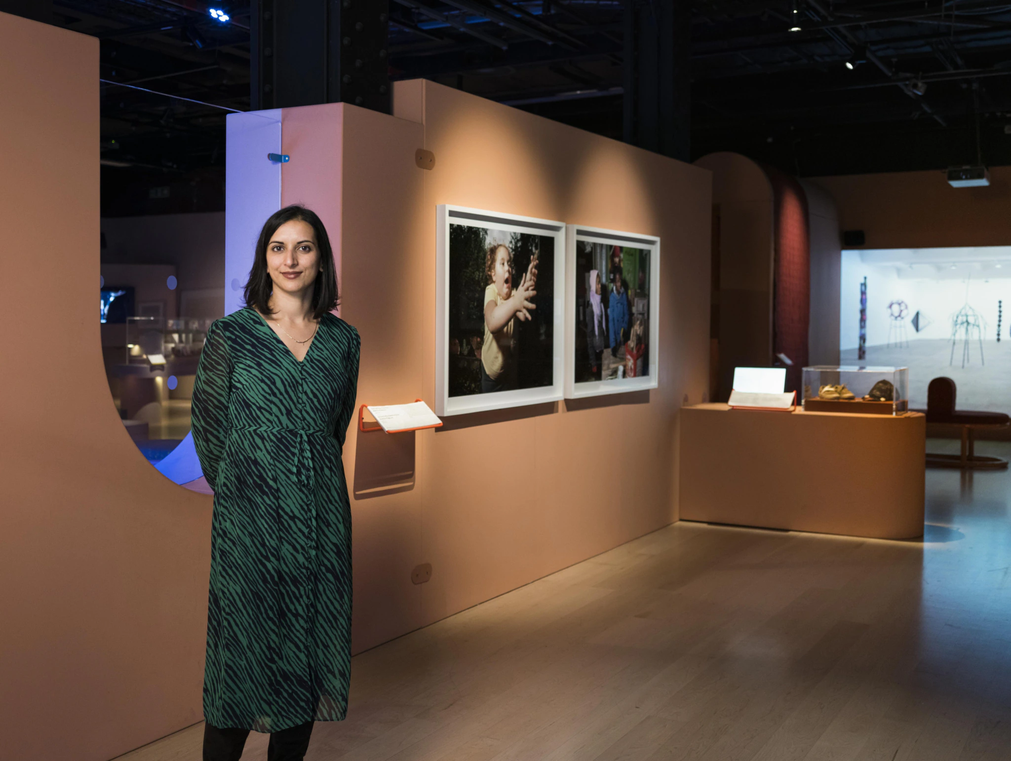 A woman in a green dress stands in a modern art gallery with light wood floors, warm-colored walls, and several framed photographs and displays visible in the background.
