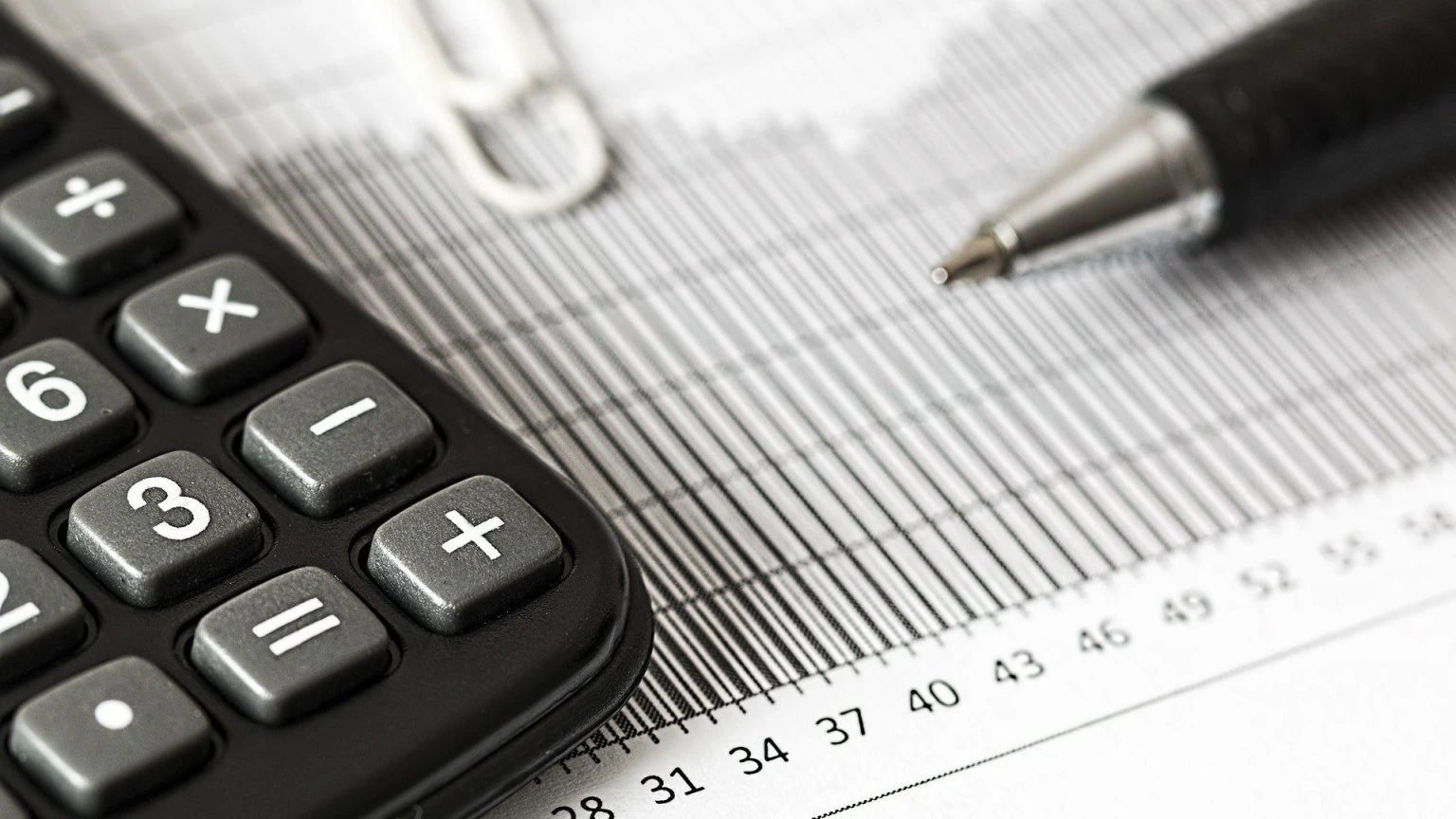 A close-up of a calculator, a pen, and a paperclip on top of a document with a graph and a ruler, suggesting financial calculations or business analysis.