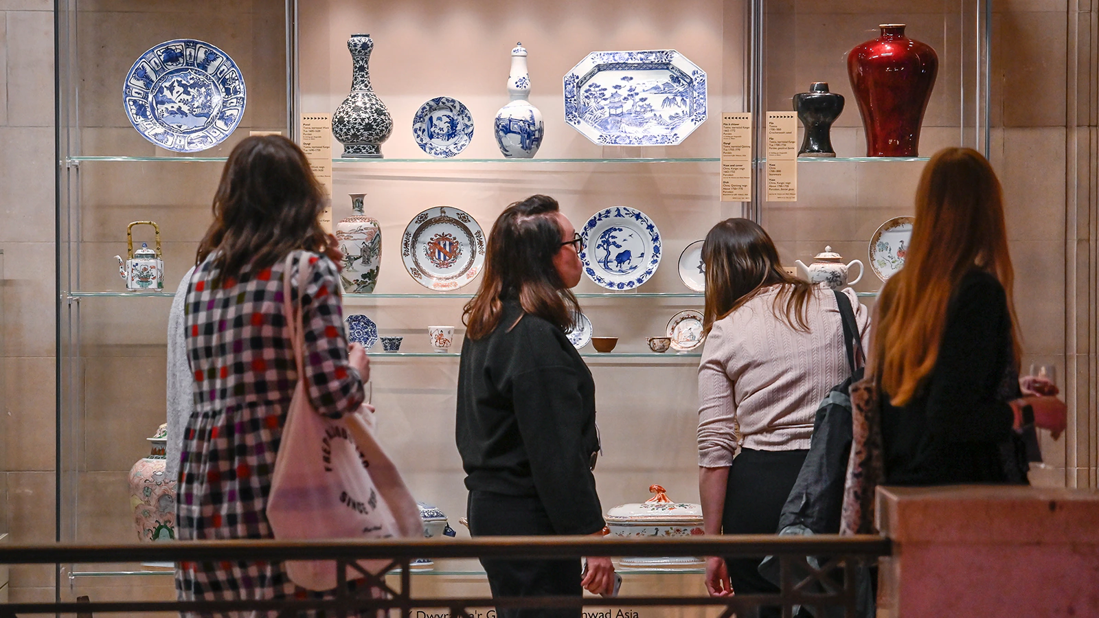 Four people stand in front of a glass display of decorative porcelain plates, vases, and ceramics in a museum or gallery setting. The display includes blue and white and red pottery pieces on glass shelves.