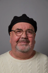 A man with glasses, a black beret, and a white cable-knit sweater smiles slightly against a plain grey background.