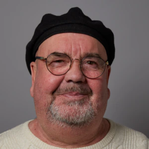 A man with glasses, a black beret, and a white cable-knit sweater smiles slightly against a plain grey background.