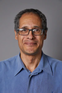 A middle-aged man with short curly hair, wearing glasses and a blue collared shirt, smiles softly against a plain gray background.