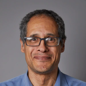 A middle-aged man with short curly hair, wearing glasses and a blue collared shirt, smiles softly against a plain gray background.