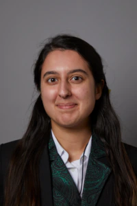 A young person with long dark hair, wearing a black jacket with a green paisley pattern and a white collared shirt, stands against a plain gray background, looking at the camera with a neutral expression.