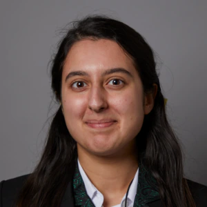 A young person with long dark hair, wearing a black jacket with a green paisley pattern and a white collared shirt, stands against a plain gray background, looking at the camera with a neutral expression.
