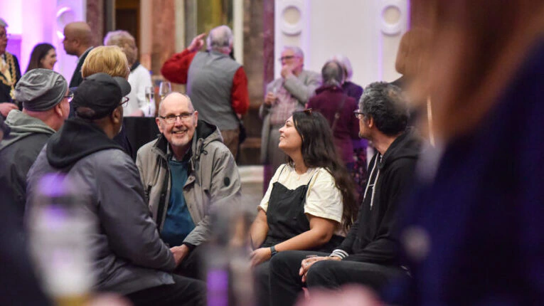 A group of people sit in a circle, talking and smiling, inside a large, elegant room with ornate columns and purple lighting. Other attendees are seen mingling in the background.