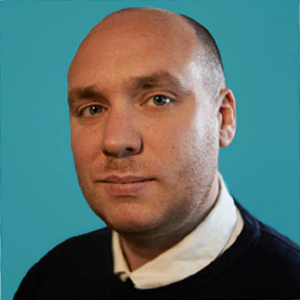 A man with a shaved head, light skin, and blue eyes wearing a dark sweater over a white collared shirt, posing in front of a solid teal background.