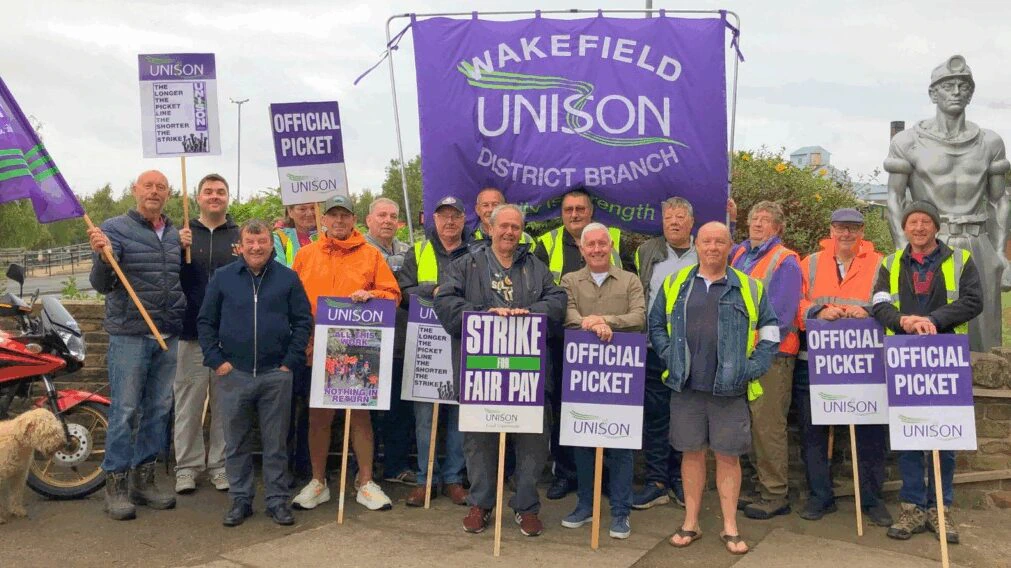 A group of people stand together holding signs that say Official Picket and Strike For Fair Pay in front of a large purple Wakefield UNISON District Branch banner. Some wear high-visibility vests.