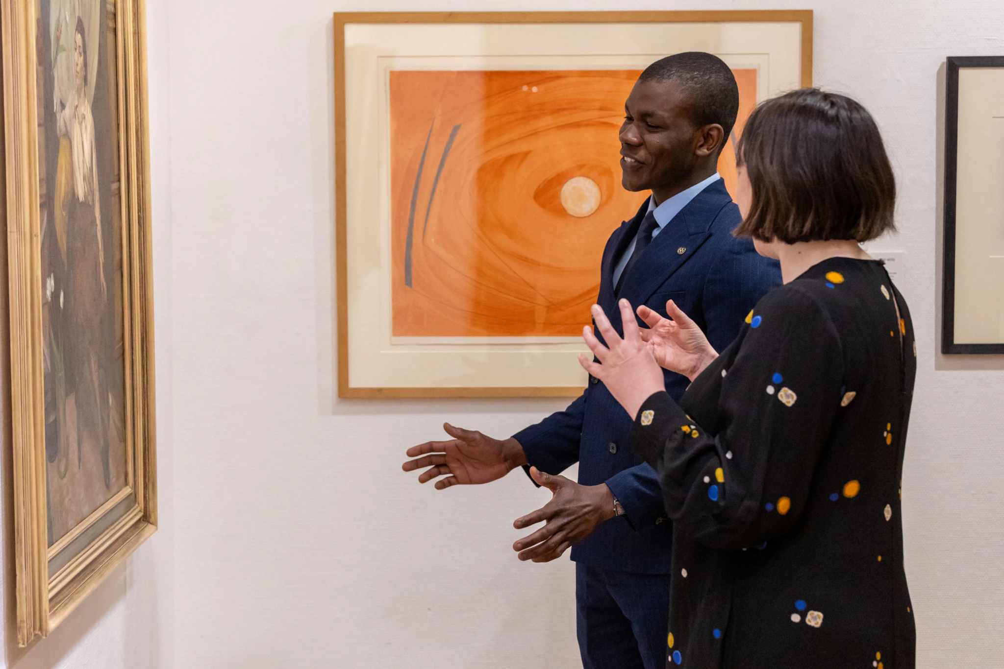 Two people stand in an art gallery, engaged in conversation while looking at a framed painting on the wall. Other abstract artworks are visible in the background.