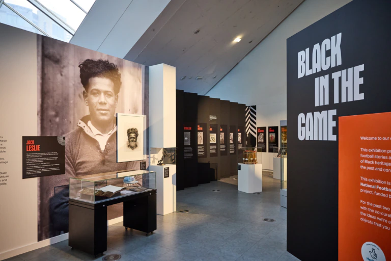 A museum exhibit titled Black in the Game features a large photograph of footballer Jack Leslie, display cases with memorabilia, and informational panels about Black football history.