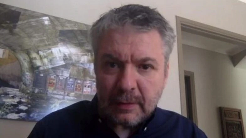 A middle-aged man with gray hair and a beard is looking at the camera in a room with light-colored walls and a painting of a tunnel behind him. He is wearing a dark shirt. A video play button is visible in the lower left corner.