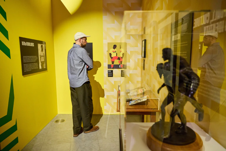 A man wearing a cap and glasses observes exhibits in a brightly lit yellow museum room, with information panels, a small table, and a dark statue displayed in a glass case.
