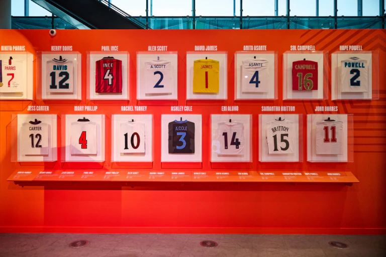 A display of framed football shirts with player names and numbers, arranged in two rows on an orange wall, each with a small description below. A large window lets in natural light above the display.