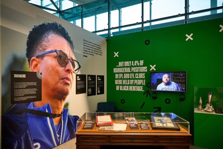 Museum exhibit featuring a large photo of a person wearing glasses, a wall with statistics about Black representation in football management, a TV screen, and a display case with documents and memorabilia.